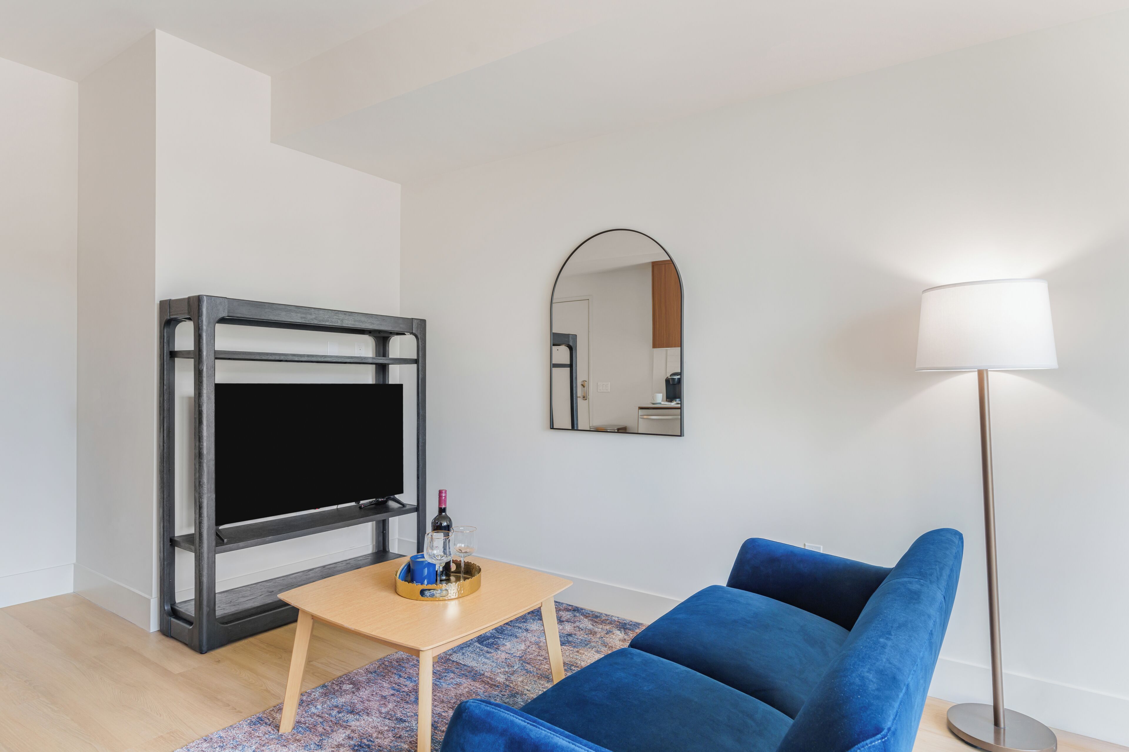 Ein modernes Wohnzimmer mit Flachbildfernseher, einem blauen Sofa, einem kleinen Holztisch, einer Stehlampe und einem Spiegel an der Wand.