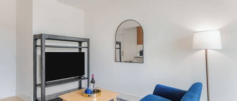 Ein modernes Wohnzimmer mit Flachbildfernseher, einem blauen Sofa, einem kleinen Holztisch, einer Stehlampe und einem Spiegel an der Wand.