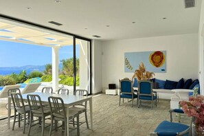 A modern interior with a dining area, a sofa, and a large window overlooking a pool and landscape. - Villa Porto Mella in Vasilikos, Zakynthos (Zakinthos)