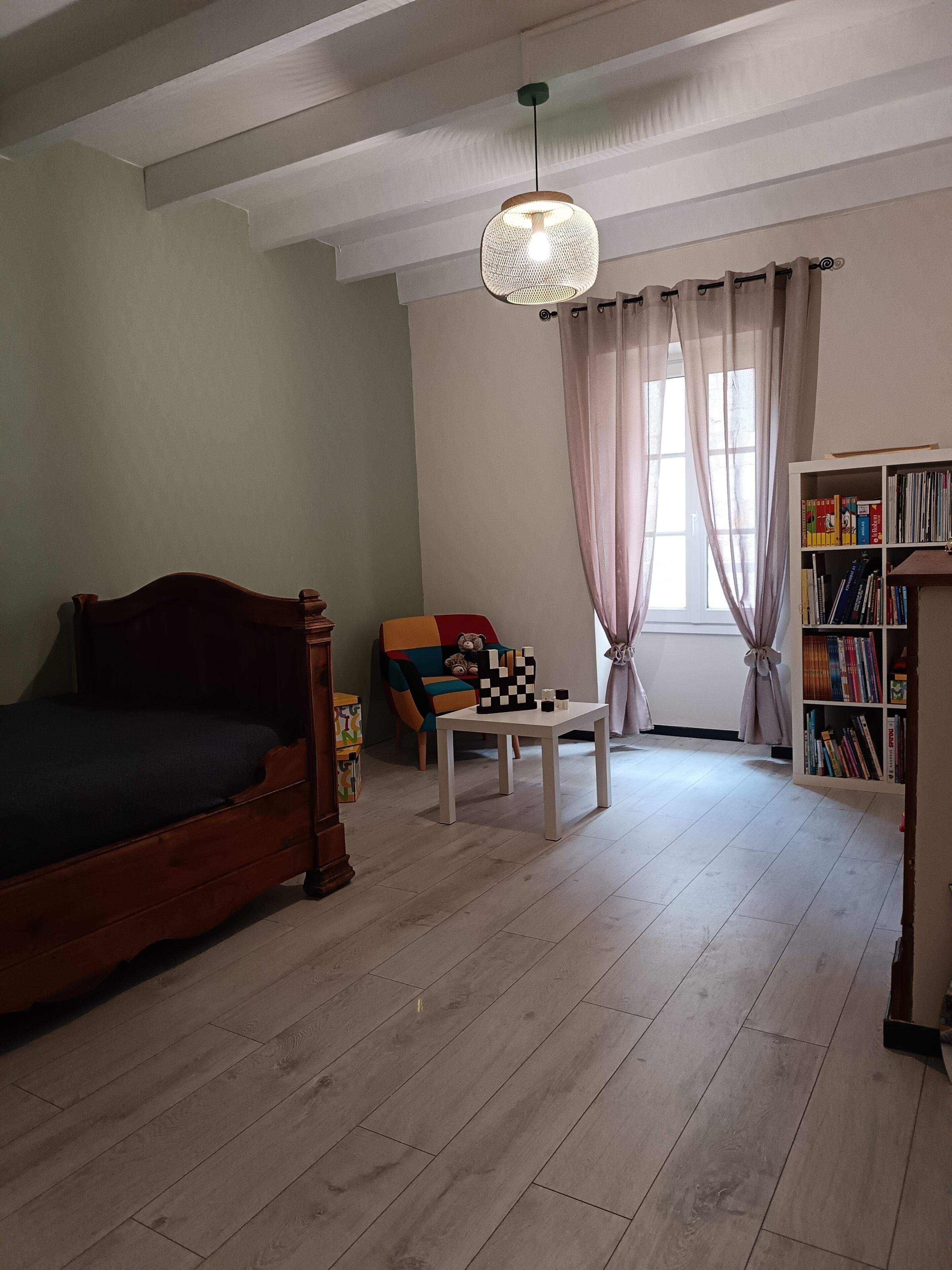 Une chambre à coucher avec un lit en bois, une petite table sur laquelle est posé un jeu d’échecs, une bibliothèque et une fenêtre avec des rideaux.
