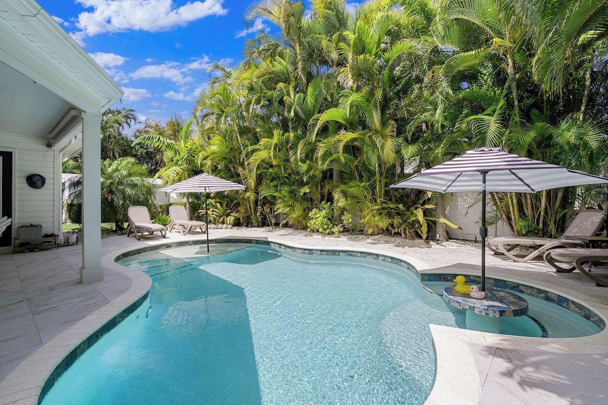 Una piscina de borde curvo, rodeada de sillones de descanso y sombrillas, con palmeras al fondo.