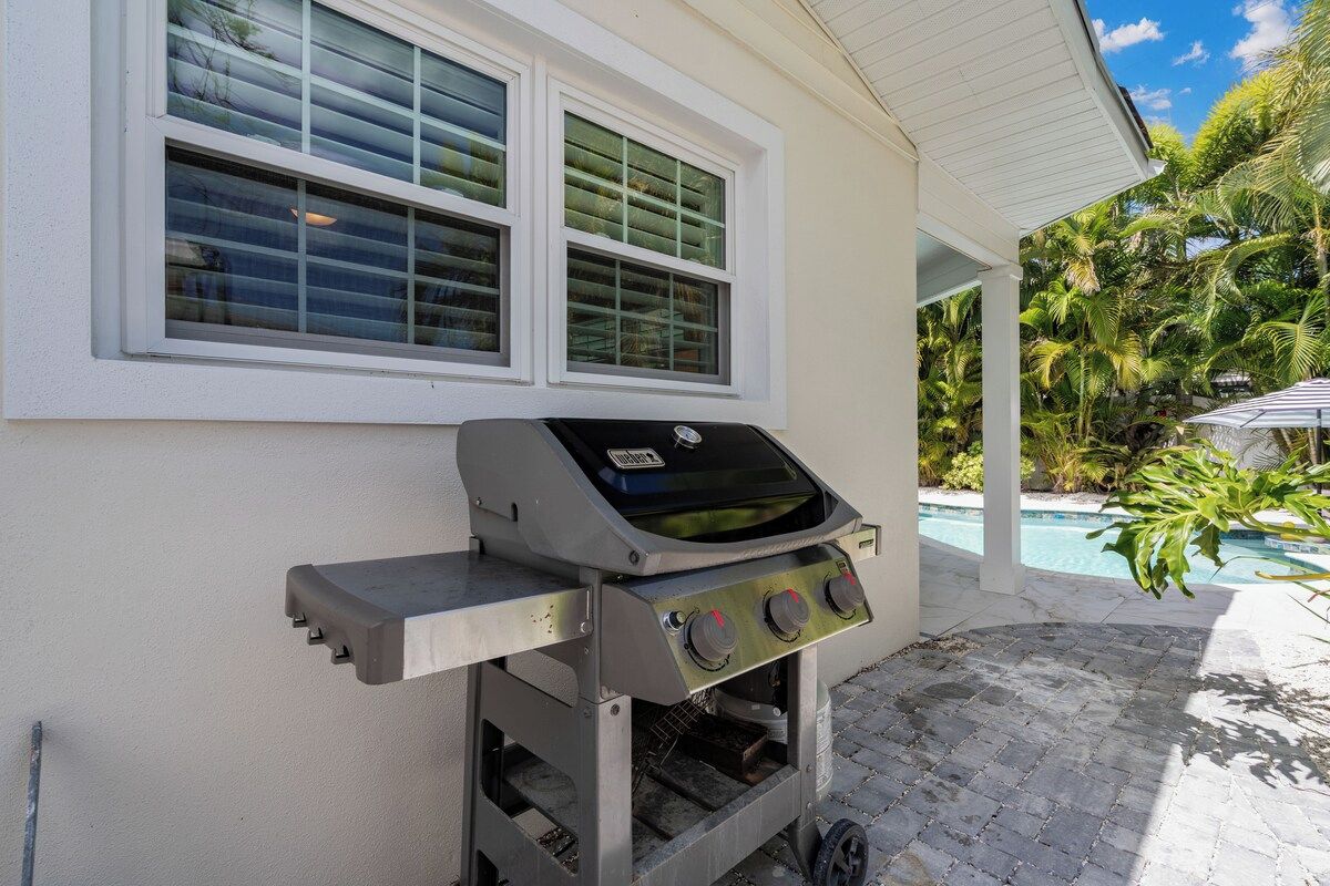 A portable gas grill with a digital display, positioned against a house wall with a window and a view of a swimming pool and tropical vegetation.