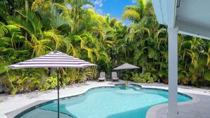 Une piscine bordée de deux chaises longues et d’un parasol à rayures, le tout entouré d’une végétation tropicale luxuriante.