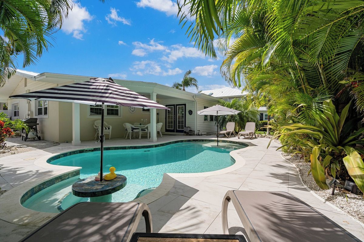 A swimming pool with lounge chairs and umbrellas, surrounded by palm trees and a covered patio area.