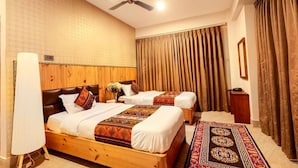 Habitación de hotel con dos camas, ventilador de techo, cabecero de madera, alfombra estampada y un gran ventanal con cortinas.
