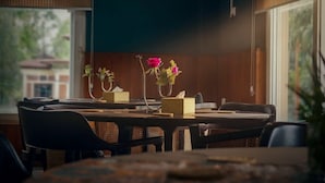 Un comedor con una mesa preparada para dos personas, un jarrón con flores y una ventana con vistas a edificios.