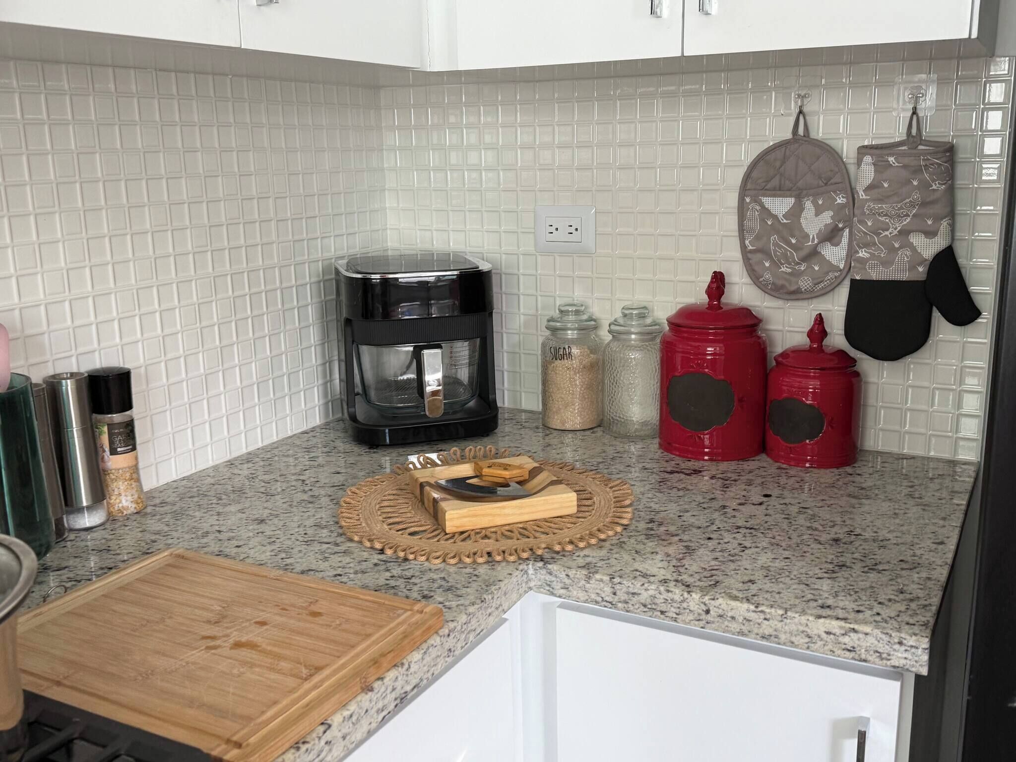 Una cocina con salpicadero de azulejos blancos, una cafetera, frascos de especias y guantes de horno colgando.