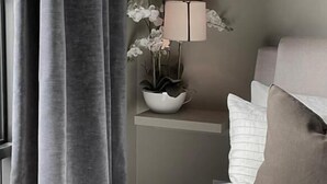 Un dormitorio con cama, almohadas, una mesita de noche con lámpara y flores, y cortinas grises.