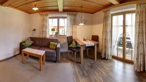 A room with a wooden ceiling, a sofa, a table, and a window with curtains. - Vacation apartment Guat'kammer (Inzell)