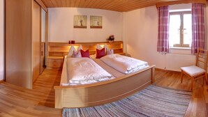A wooden bed with two pillows, a nightstand, a chair, and a window with curtains. - Vacation apartment Guat'kammer (Inzell)