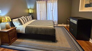 Una camera da letto con un letto, una ventola a soffitto, un comodino, una lampada, una cassettiera e una finestra con tende.