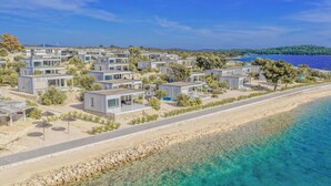 Una zona costiera con unità abitative moderne e compatte disposte lungo una spiaggia sabbiosa, con acque blu cristalline e vista panoramica su un paesaggio collinare.