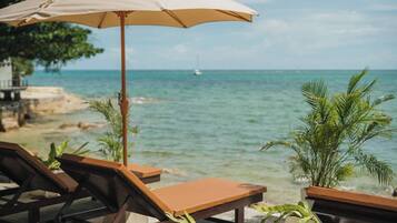 A beachside lounge with wooden chairs and a beige umbrella, overlooking a calm sea and a distant sailboat.