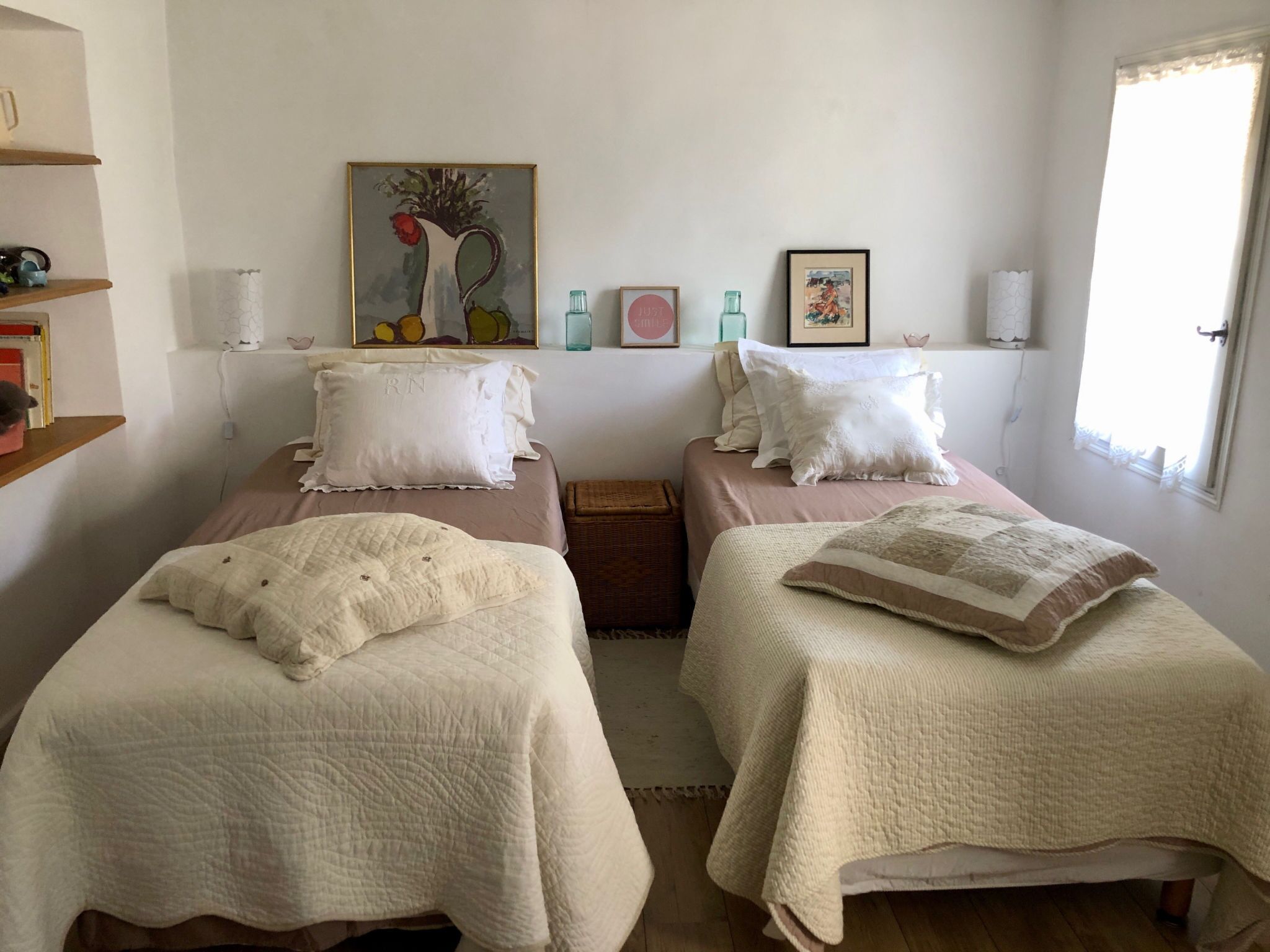 Un dormitorio con dos camas, cada una con colcha blanca acolchada y almohadas, una pequeña cesta de mimbre y un cuadro sobre las camas.