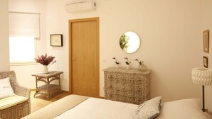 A bedroom with a bed, a wicker chair, a small table with a vase, a dresser, a mirror, and a door.