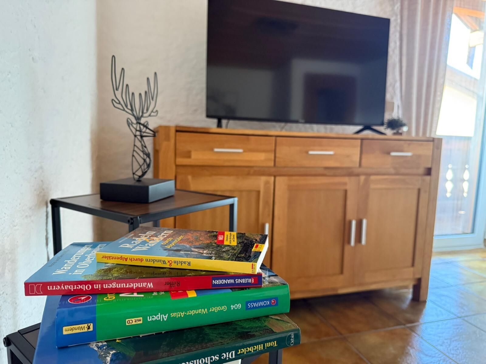 A stack of books on a table with a TV and wooden cabinet in the background.