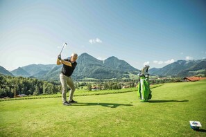 A golf course with a golfer in mid-swing, a golf bag, and a distant mountain range. - Vacation apartment BergZeit for max. 4 adults (Chieming)