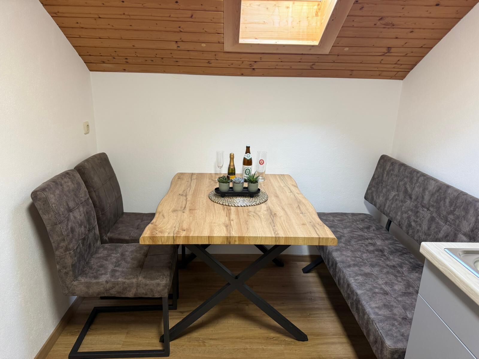 Un comedor con una mesa de madera, dos bancos grises tapizados y una pequeña zona de cocina con un fregadero.