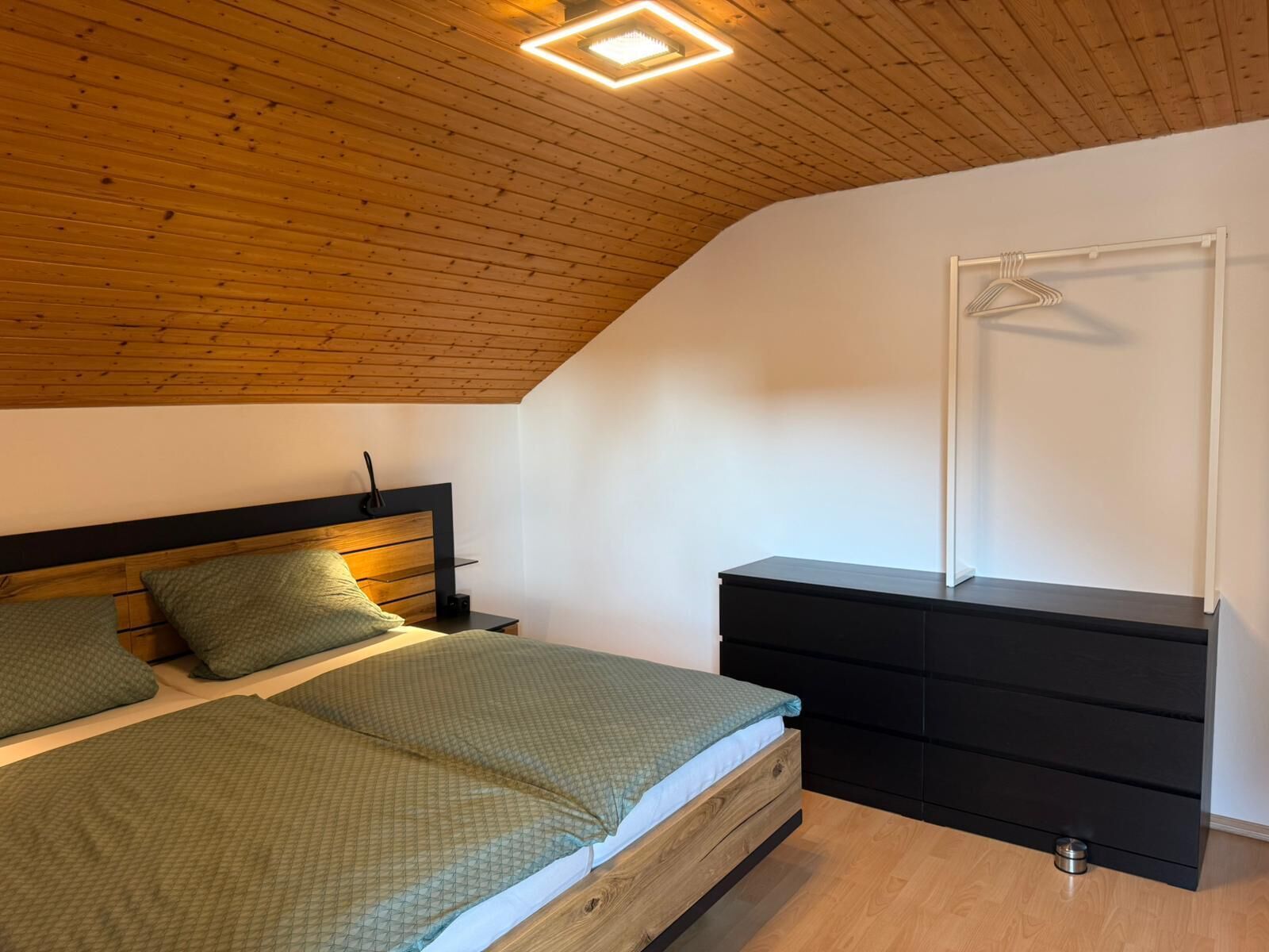 Une chambre avec un plafond en pente, un lit avec une literie verte, une commode noire et une armoire blanche avec un cintre.
