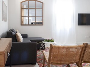 A modern living room with a sofa, a small round table with flowers, a wicker chair, a large mirror, and a TV mounted on the wall. - Apartamento en Puerta del Sol centro ciudad (Madrid)