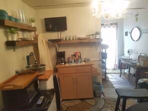 A compact kitchen with a TV, shelves with dishes, a coffee maker, and a dining area with a table and chairs. - Adorable tiny house near Mercedes Benz Stadium (Atlanta)