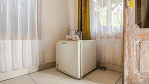 Ein kleiner, weißer Kühlschrank mit Wasserspender, ein Wasserkocher und ein Teller mit einer Tasse in einer Ecke eines Raumes mit weißen Wänden und einem Fenster mit durchsichtigen Vorhängen.