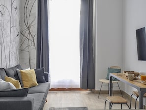 A modern living room with a grey sofa, a dining table with chairs, a flat-screen TV, and a large window with curtains. - Apartamento en Puerta del Sol centro ciudad (Madrid)