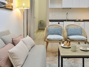 Una sala moderna con un sofá, dos sillas de mimbre, una mesa de centro y una cocina con armarios blancos y un lavabo.