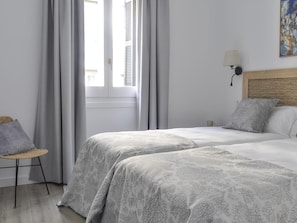 Habitación de hotel con dos camas, una silla de mimbre, una lámpara y un cuadro en la pared.