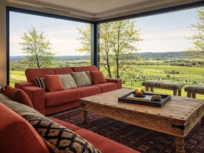 Ein modernes Wohnzimmer mit einem roten Sofa, einem Holztisch und Blick auf eine grüne Landschaft durch große Fenster.