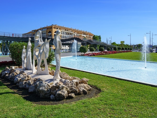 Une fontaine ornée de sculptures, située dans un espace paysager, un pont et un bâtiment en arrière-plan.