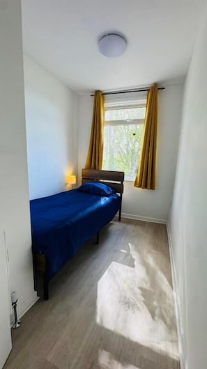 Una habitación pequeña con cama individual, ventana, lámpara de noche y luz de techo.