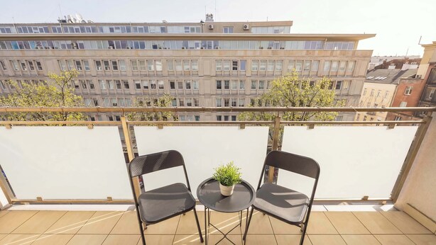 A balcony with two chairs and a small table, a potted plant, and a view of a multi-story building. - Stylish Apartment with Balcony | Center of Warsaw (Warszawa)