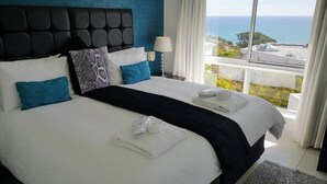 Una cama grande con sábanas blancas, cabecera negra y almohadas azules. Se ve una ventana con vista al océano y edificios.
