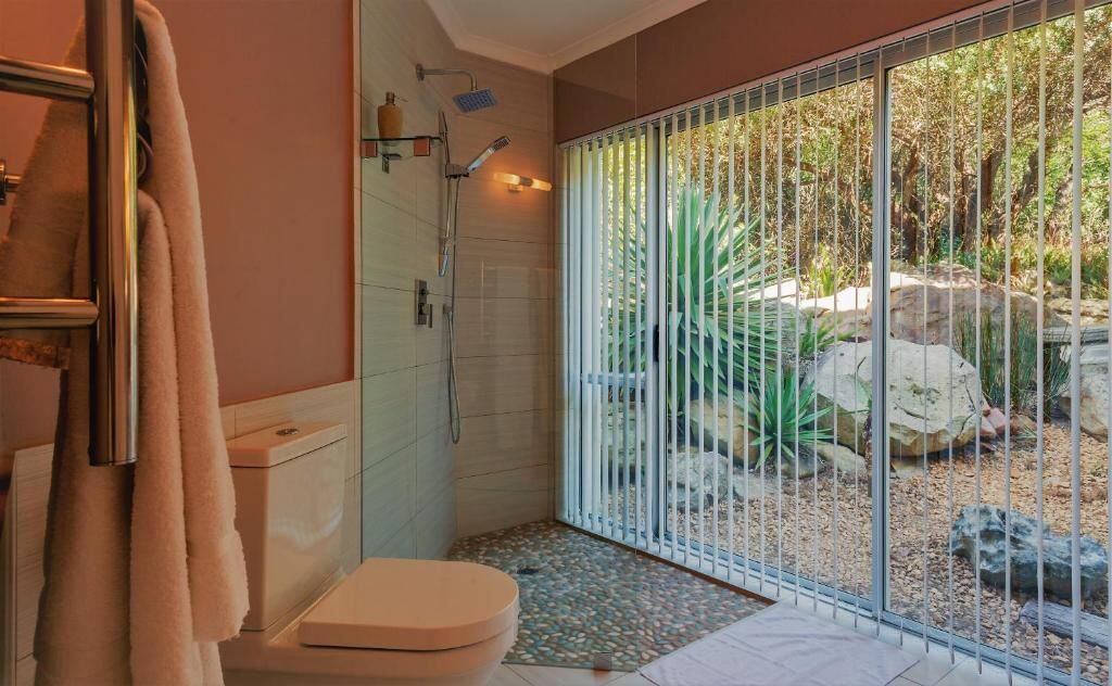 A bathroom with a toilet, shower, and a glass door leading to an outdoor area with rocks and plants.