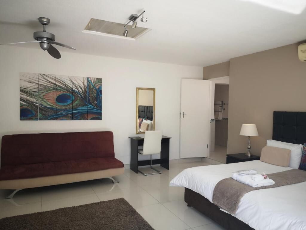 A hotel room with a bed, a desk, a chair, a red sofa, a peacock feather painting, a ceiling fan, and a door leading to a bathroom.