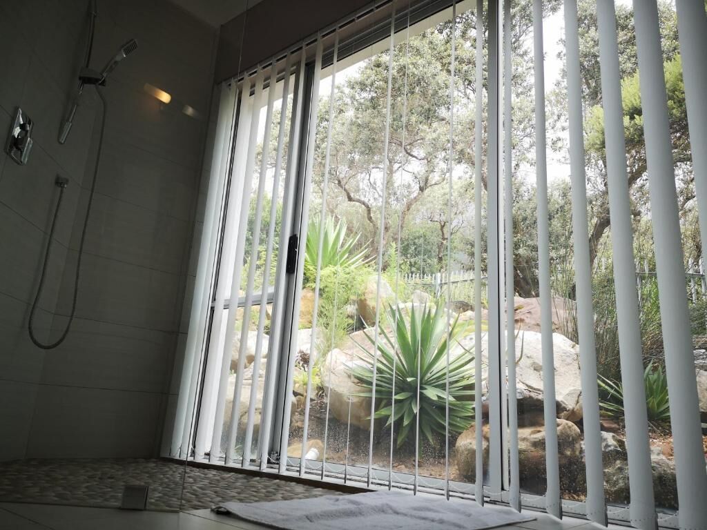 Casa de banho com chuveiro, janela com persianas verticais e vista para um jardim com pedras e plantas.
