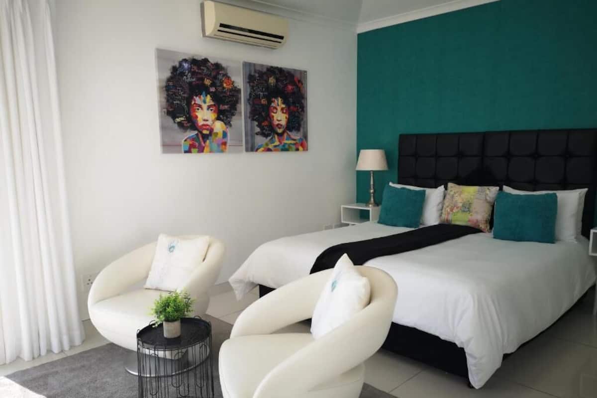 Une chambre d’hôtel moderne comprenant un lit, deux fauteuils blancs, une petite table ronde avec une plante et deux unités de climatisation fixées au mur.