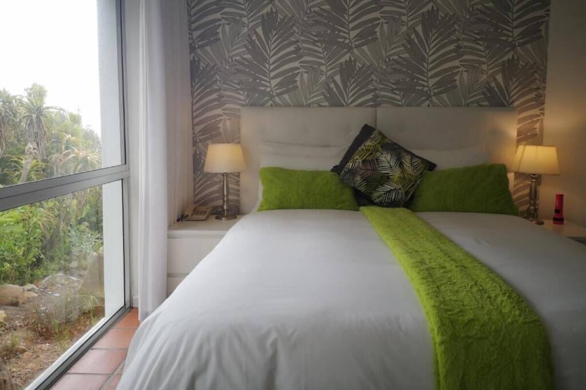 Une chambre à coucher avec un grand lit, deux lampes de chevet, une fenêtre donnant sur un paysage verdoyant et un papier peint à motifs.