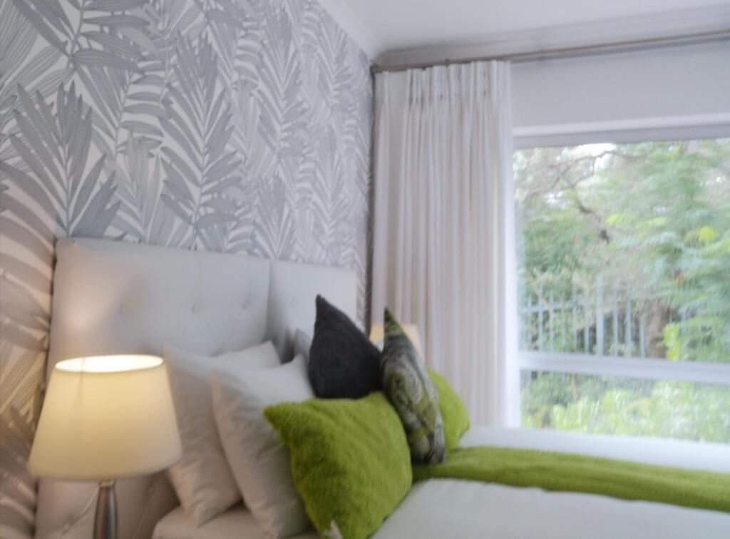 Un dormitorio con cama, almohadas, lámpara y ventana con vista a follaje.