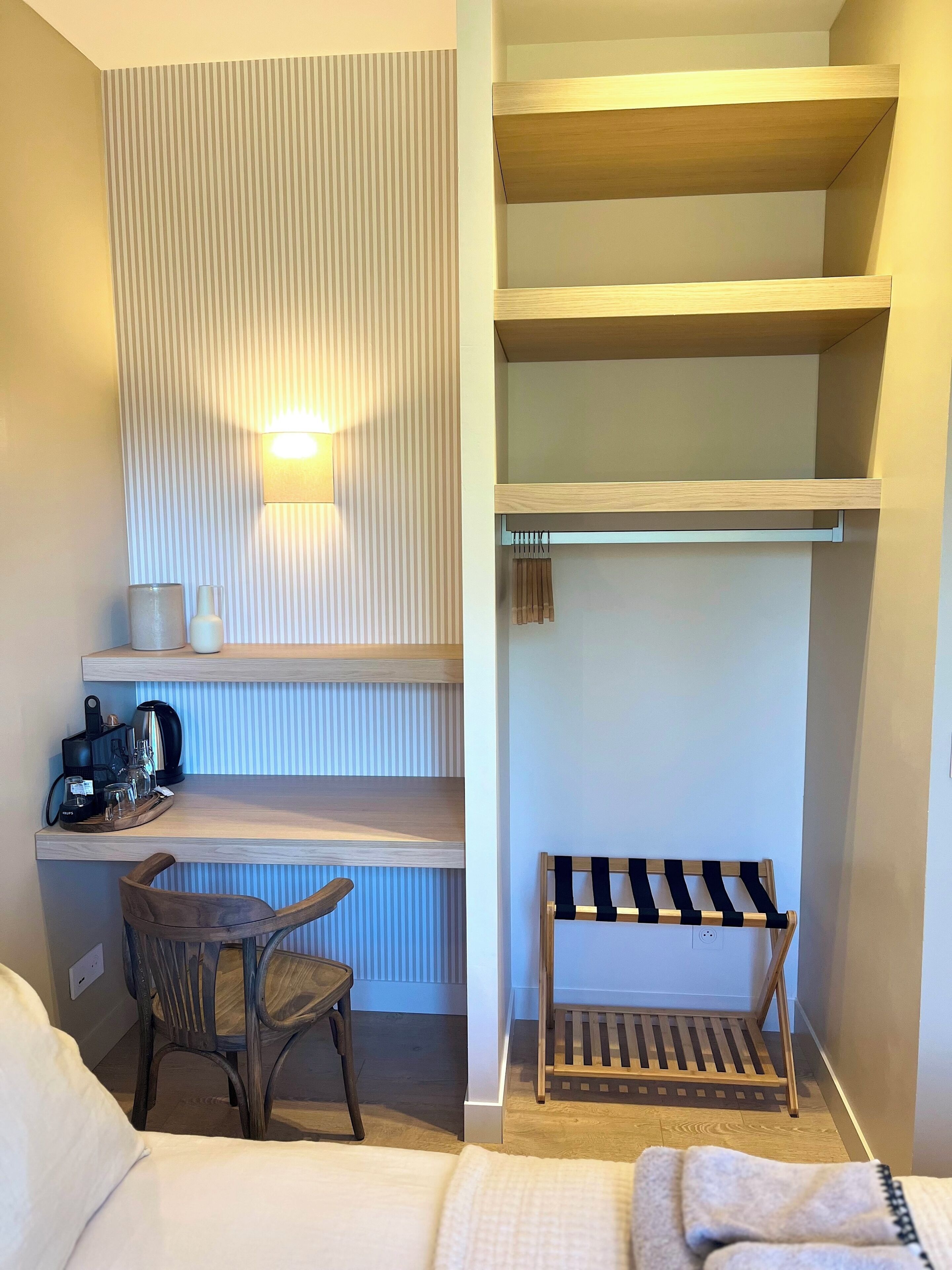 A compact room with a bed, a wooden chair, a small table with a coffee maker, and a wall-mounted shelf with hanging clothes.