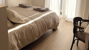 Un dormitorio con una cama, un escritorio, una silla, un televisor montado en la pared y una ventana con cortinas.