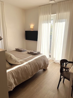 Un dormitorio con cama, escritorio, silla, televisor montado en la pared y ventana con cortinas.