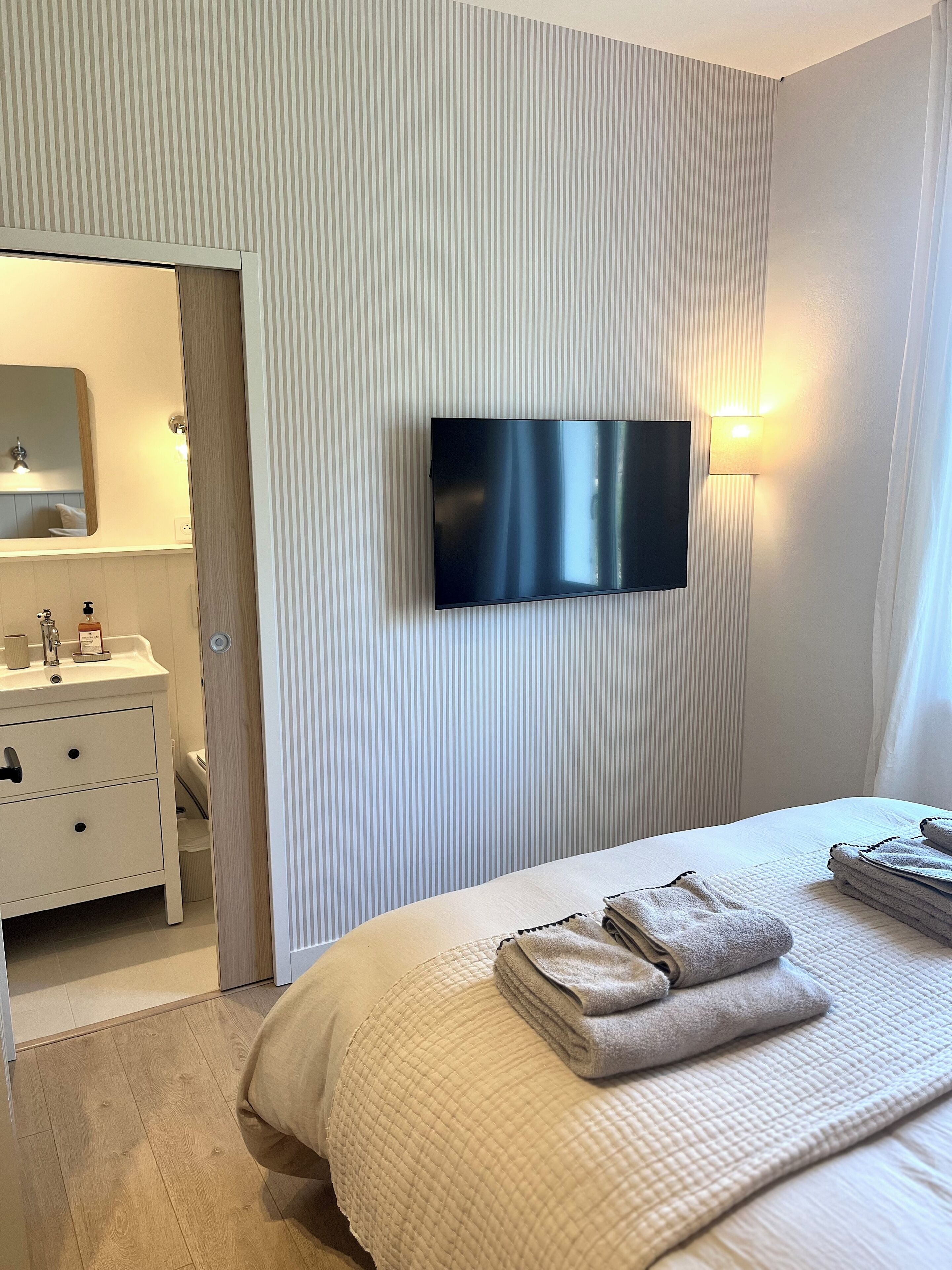 Une chambre d’hôtel moderne dotée d’un téléviseur à écran plat fixé au mur, d’une table de chevet avec une lampe et d’un lit soigneusement fait, agrémenté de serviettes.