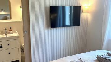 Une chambre d’hôtel moderne dotée d’un téléviseur à écran plat fixé au mur, d’une table de chevet avec une lampe et d’un lit soigneusement fait, agrémenté de serviettes.
