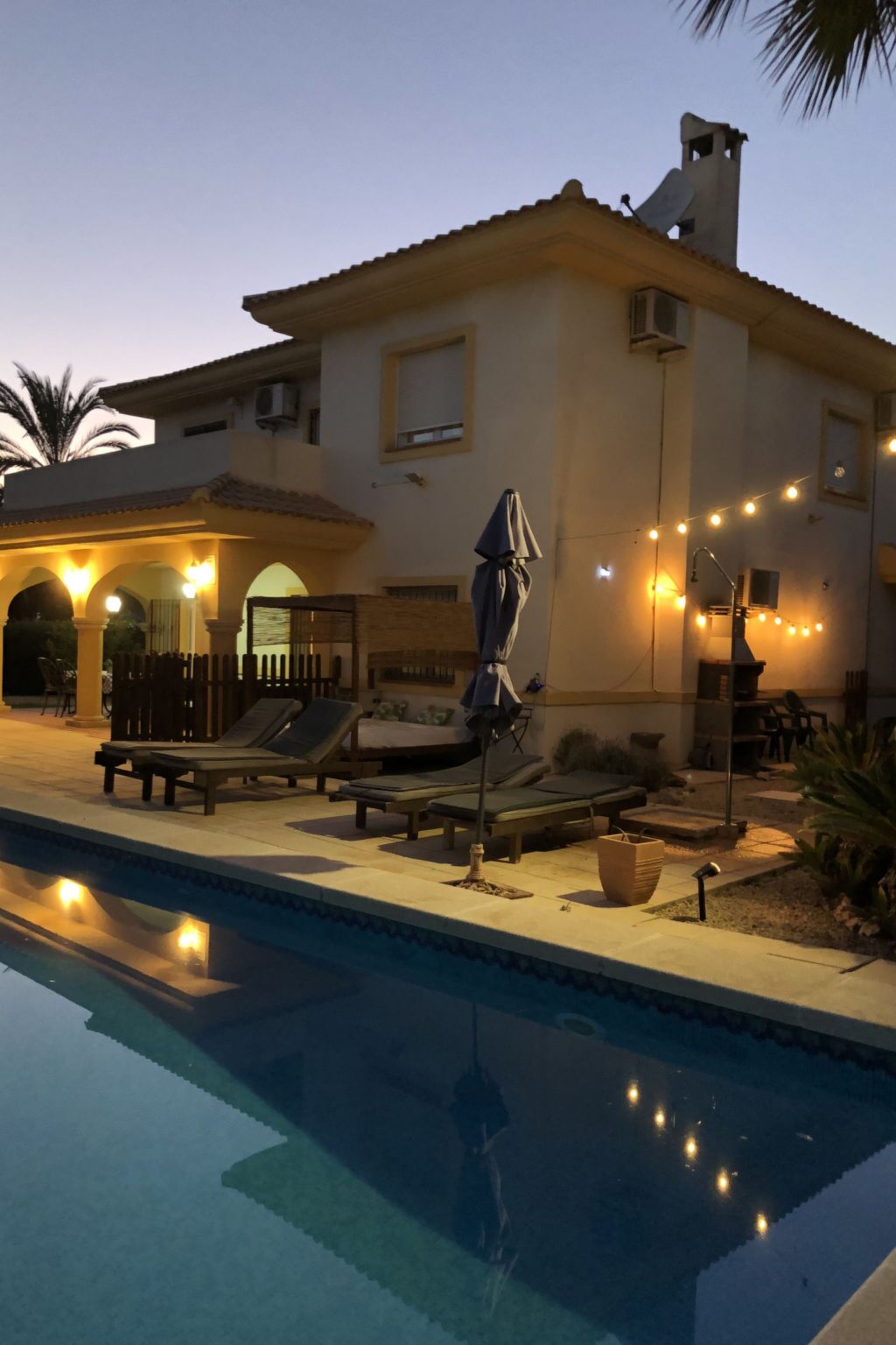 A poolside area with lounge chairs and umbrellas, a clear pool, and string lights.