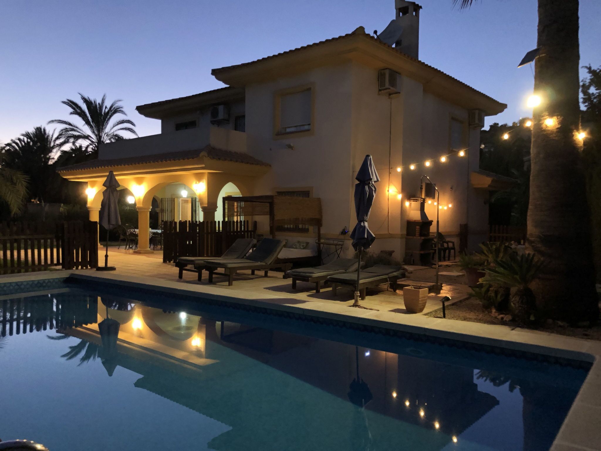 A poolside area with lounge chairs and umbrellas, a house with a balcony, and string lights.