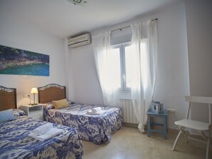 Ein Zimmer mit zwei Betten, einem Fenster mit Vorhängen, einem Heizkörper, einem kleinen Tisch mit einer Lampe und einem Sessel.