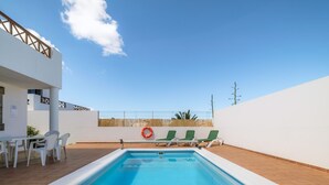 Ein rechteckiger Swimmingpool mit weißem Rand, umgeben von einer weißen Mauer und einem Holenzaun. Drei grüne Liegestühle und ein roter Rettungsboy sind vorhanden.