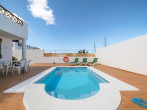 Ein rechteckiger Swimmingpool mit weißem Rand, umgeben von einer weißen Mauer und einem Holenzaun. Drei grüne Liegestühle und ein roter Rettungsboy sind vorhanden.